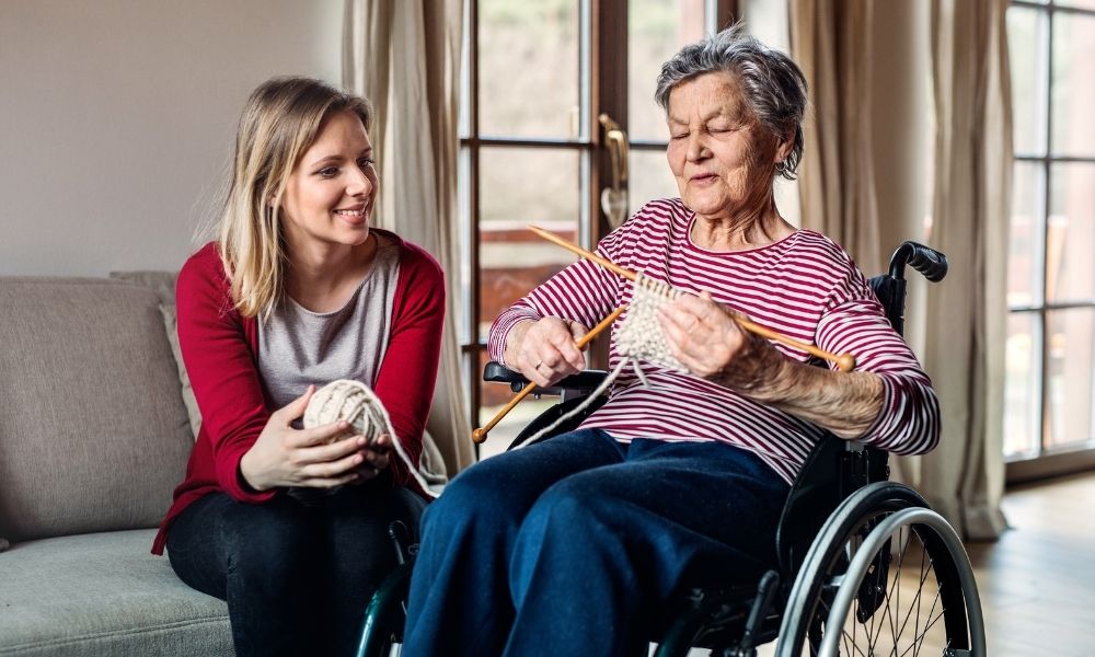 A companion talks with an older woman who is knitting in a wheelchair.