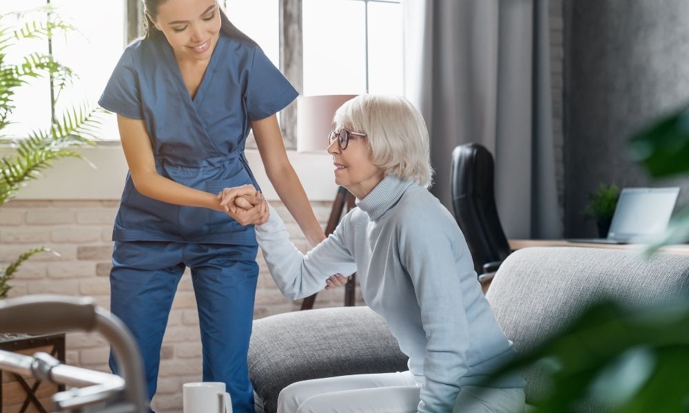 A caregiver helps an older woman rise from a sofa.