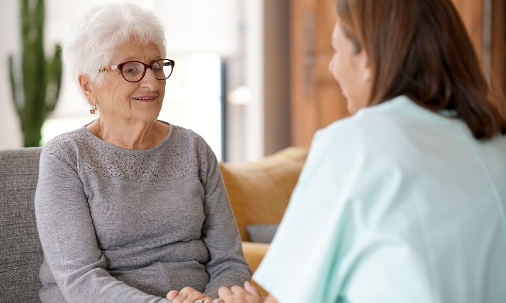 A caregiver listens to an older woman seated at home.