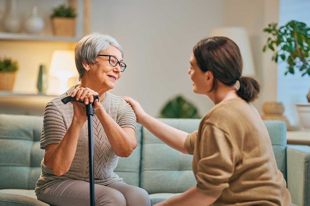 A caregiver talks with an older woman seated on a sofa.