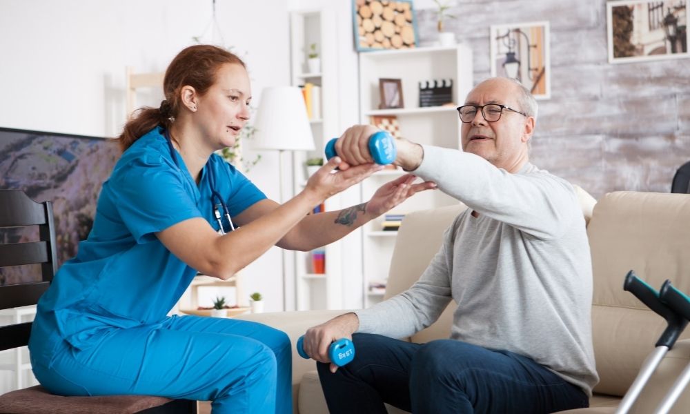 A caregiver guides an older man through a seated exercise with hand weights.
