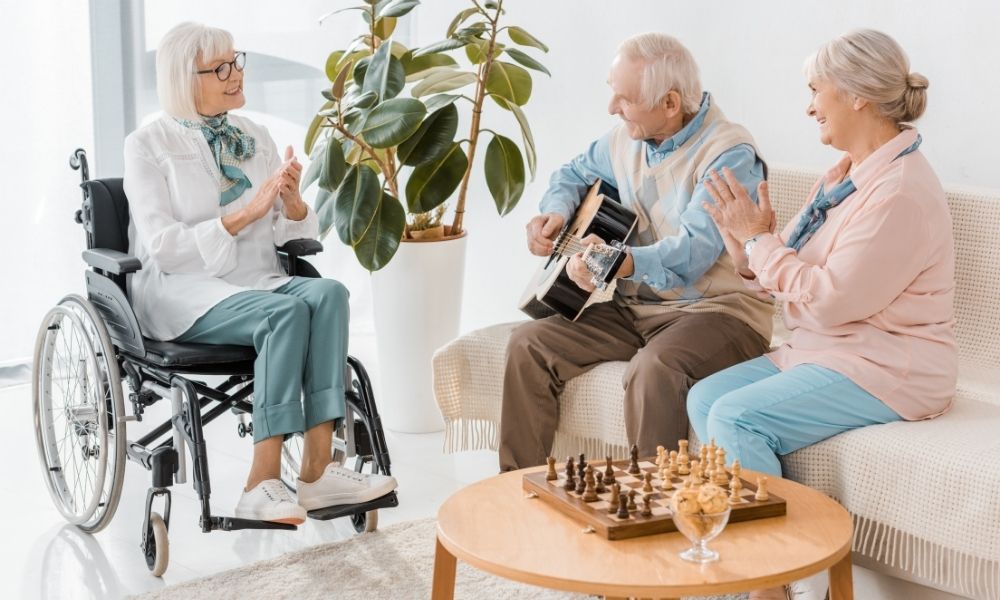 Three older adults socialize in a living room with music and a board game.