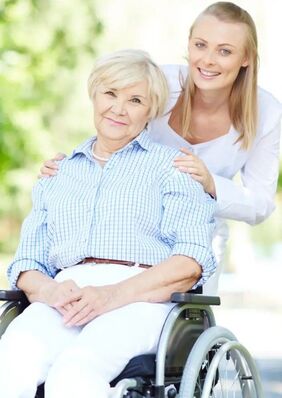 A caregiver stands behind an older woman seated in a wheelchair.